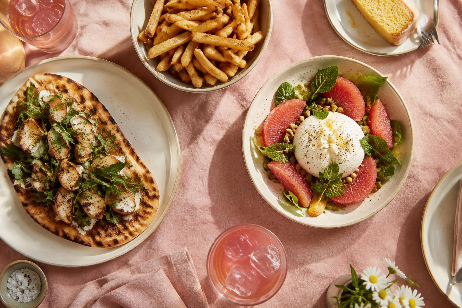 A table of flatbread, fries, burrata salad, lemon cake and pink sodas